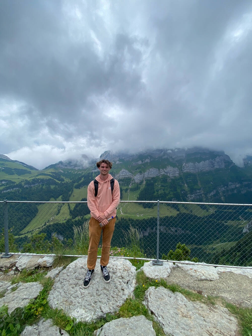 Grant Gordon standing on a mountain side in Switzerland in July of 2024 while wearing a peach sweatshirt and khaki pants with a cloudy sky.